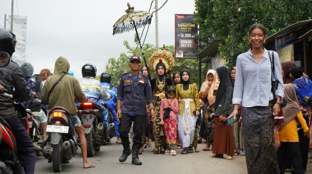 A Sasak bridal procession moves through village traffic, blending tradition and daily life in Lombok, Indonesia. A woman smiles at the camera in the foreground.