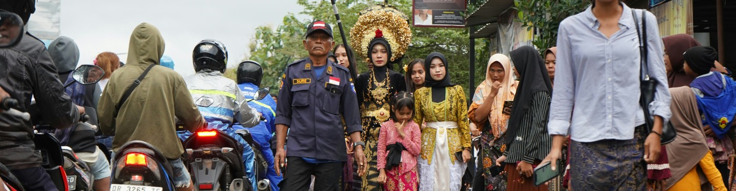 A Sasak bridal procession moves through village traffic, blending tradition and daily life in Lombok, Indonesia. A woman smiles at the camera in the foreground.